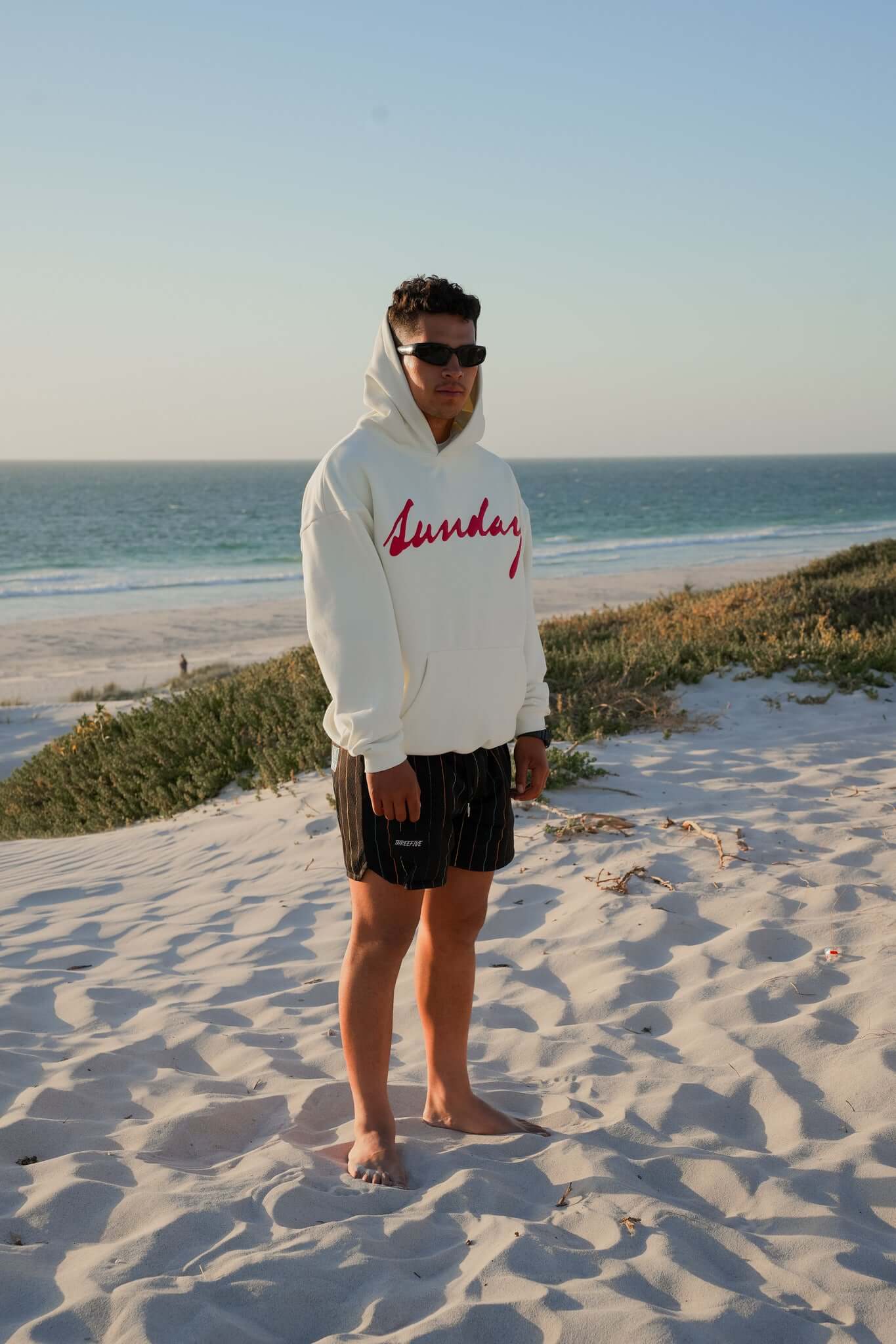 Male wearing sunglasses and vanilla coloured hoodie, standing, admiring the sunset at a beach