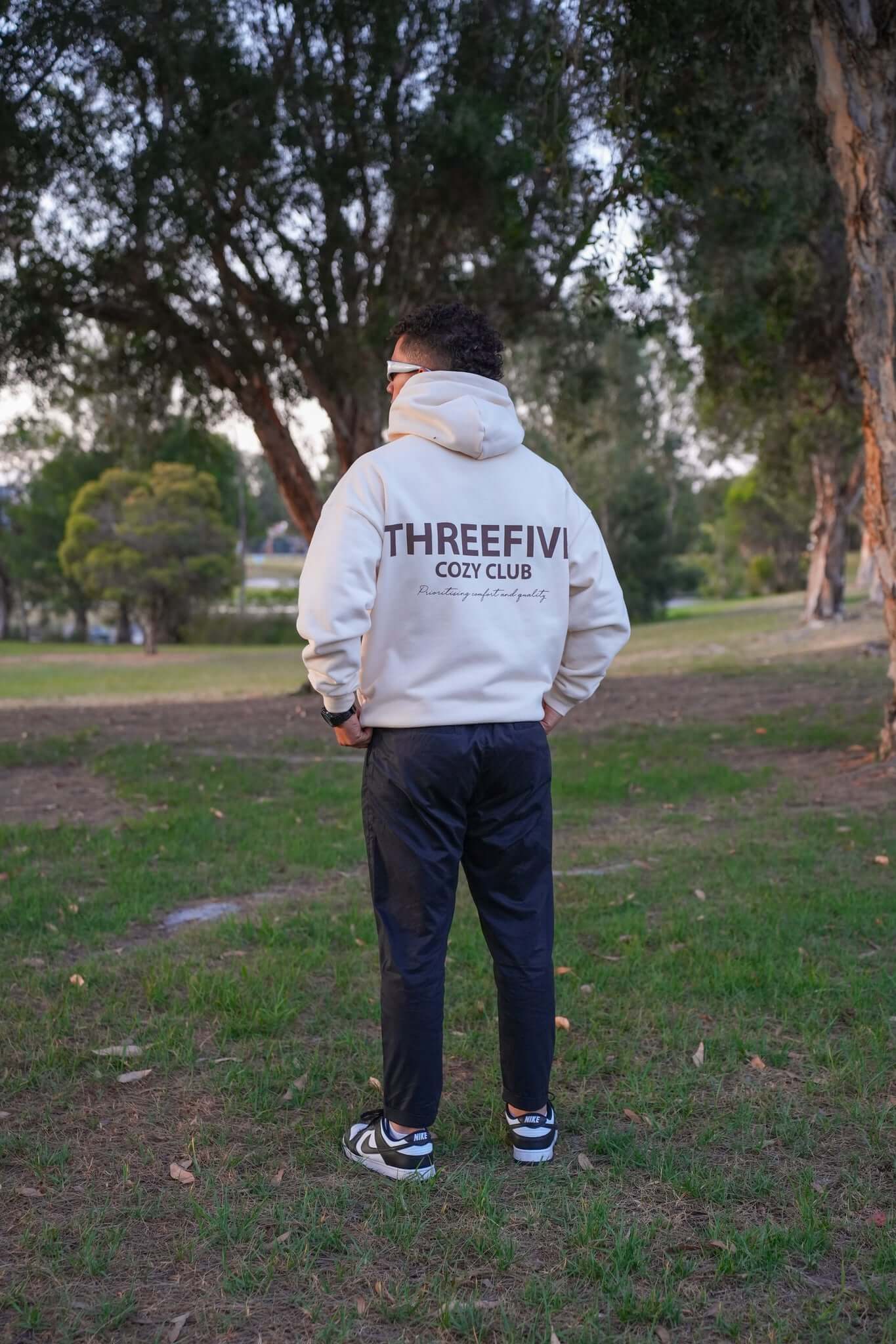 Male wearing sunglasses and a oversized cream hoodie from ThreeFive Clothing, standing confidently amidst the serene beauty of a park