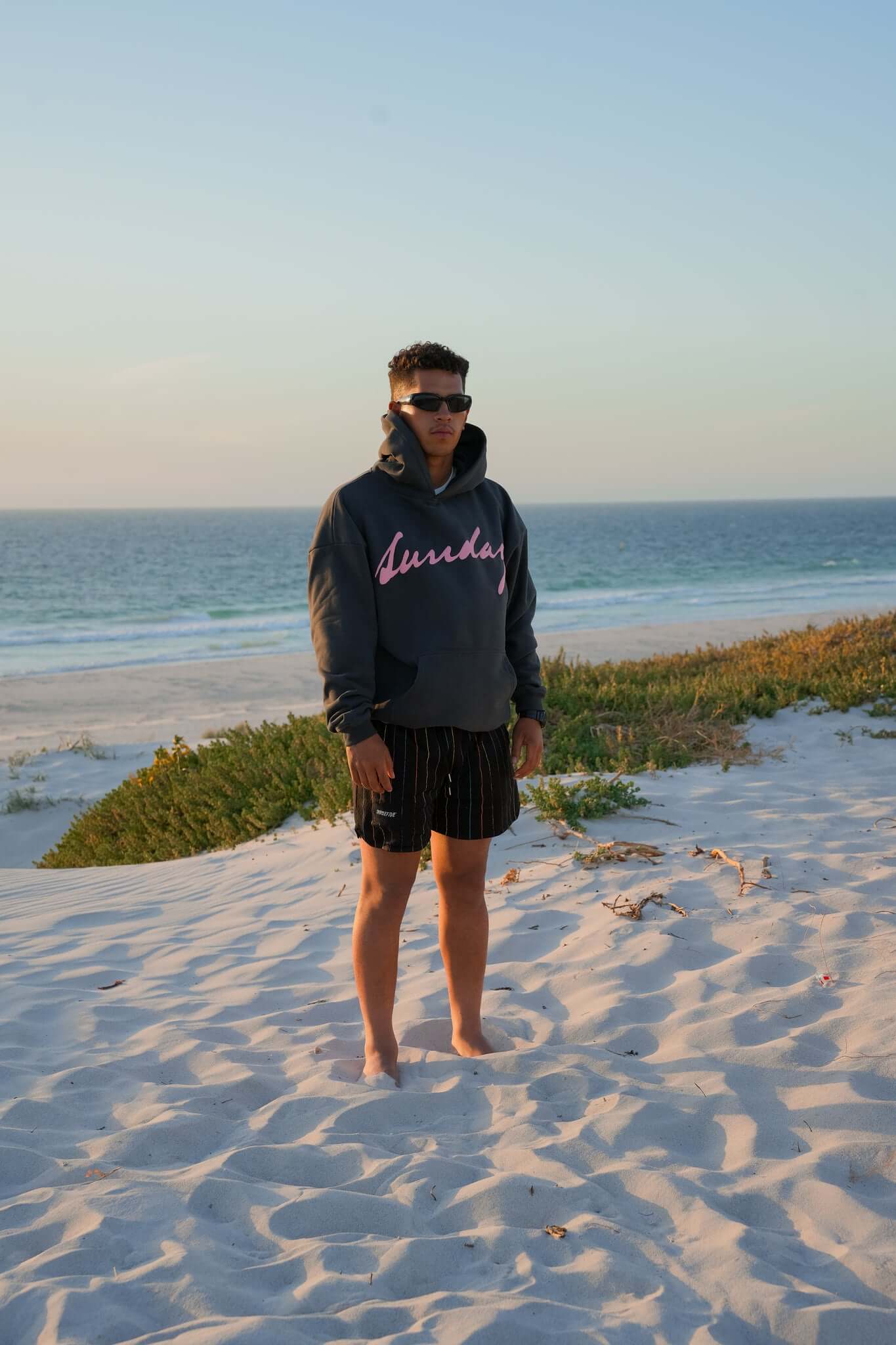 Male wearing sunglasses and a stone coloured hoodie, standing, admiring the sunset at a beach