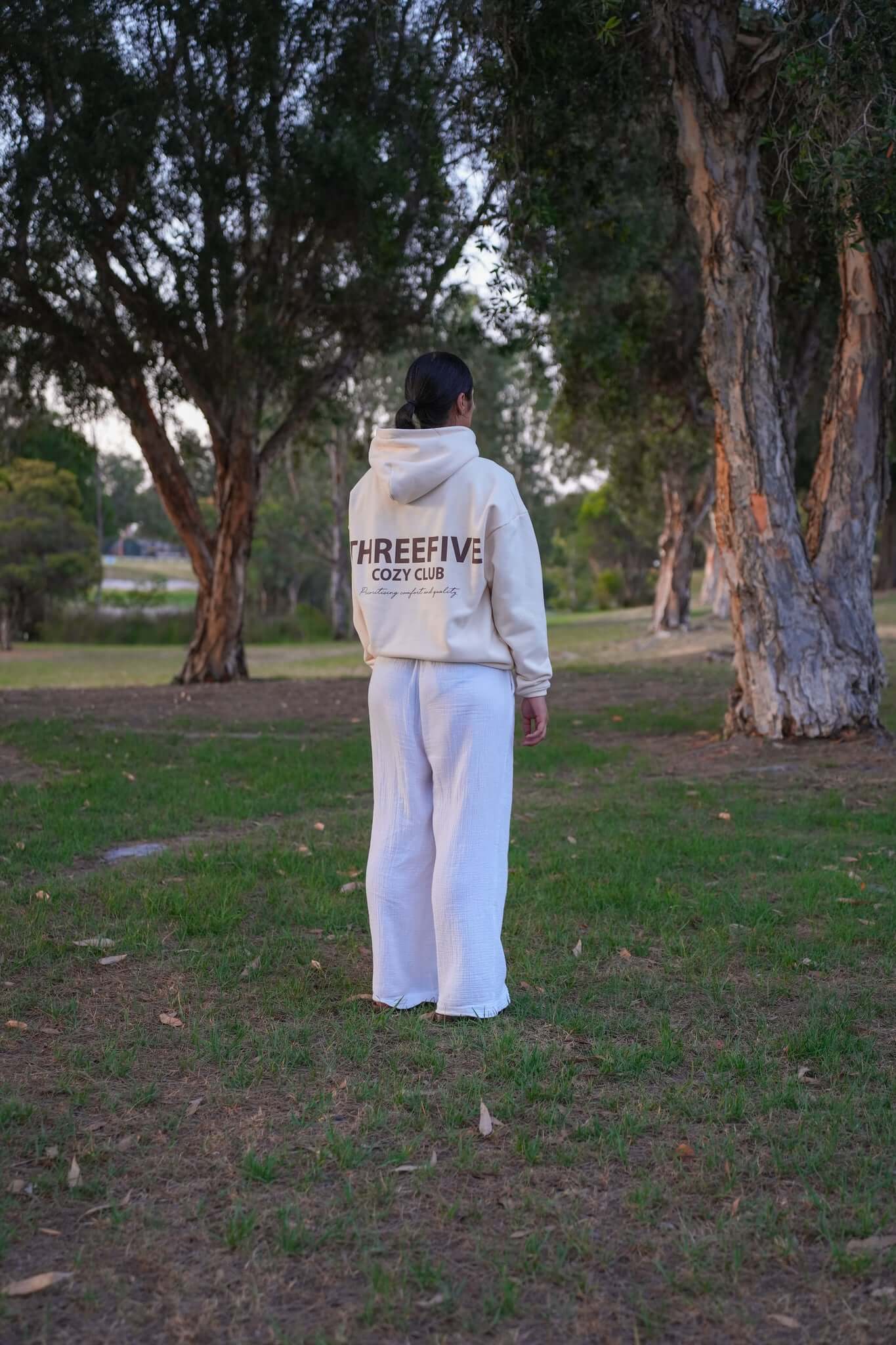 Female wearing a oversized cream hoodie from ThreeFive Clothing, facing the serene beauty of a park