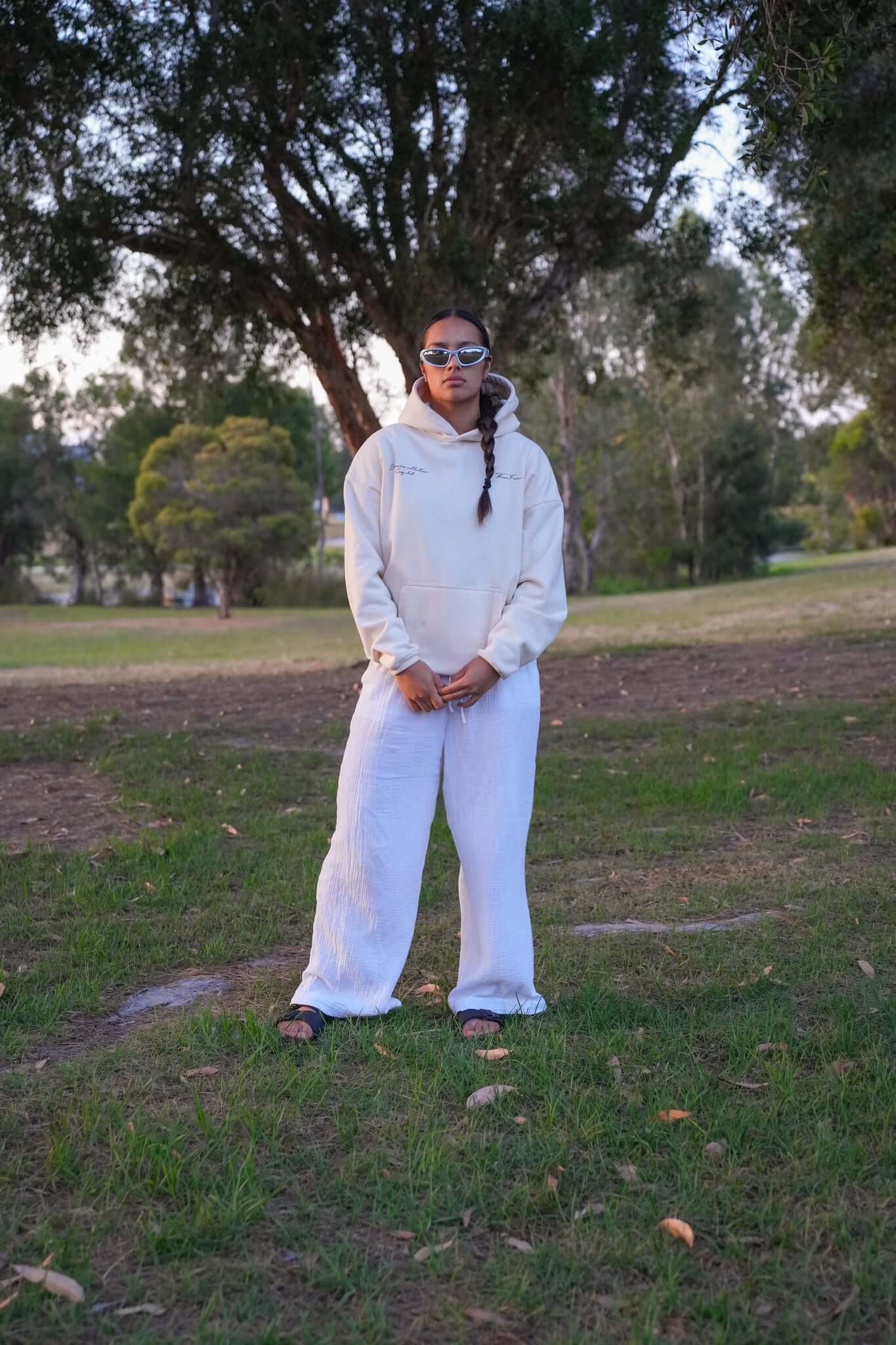 Female wearing sunglasses and a oversized cream hoodie from ThreeFive Clothing, standing confidently amidst the serene beauty of a park